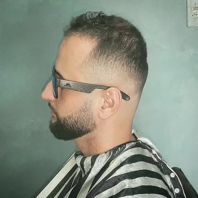 Homem de óculos escuros com degradê baixo e barba alinhada, de perfil, com a capa listrada da barbearia
