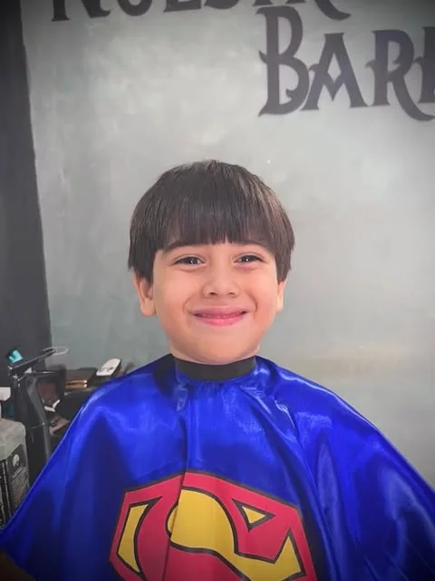 Menino sorrindo na cadeira com uma capa do Super-Homem, diante da parede com o nome Nuestra Barba pintado